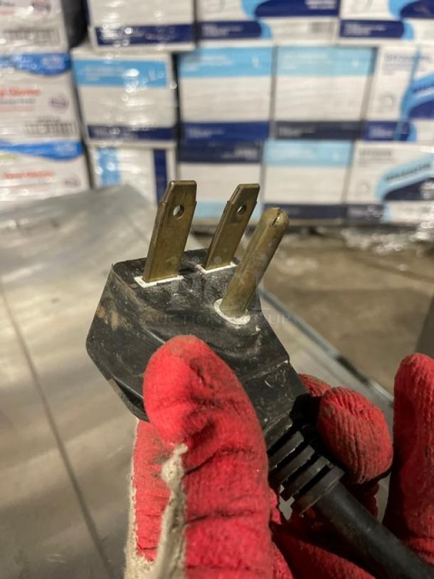 Close-up of a three-pronged plug held by a gloved hand. Background shows stacked boxed items.