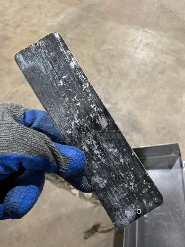 A worn metal tag held in a gloved hand, partially obscured text, surrounded by a stainless steel background.