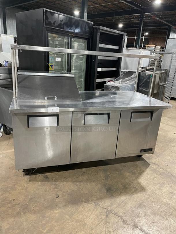 True TSSU-72-12M-B 3-door stainless steel refrigerated prep table with overhead shelf and wired rack, on casters.