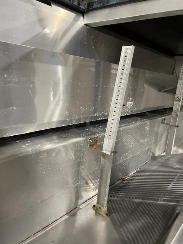 True TSSU-72-12M-B refrigerated prep table, stainless steel, 3 doors, overhead shelf, wired rack, on casters, visible wear.