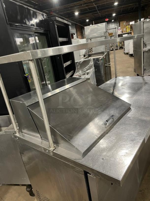 True TSSU-72-12M-B refrigerated prep table, 3 doors, stainless steel, with overhead shelf and wired rack, on casters.