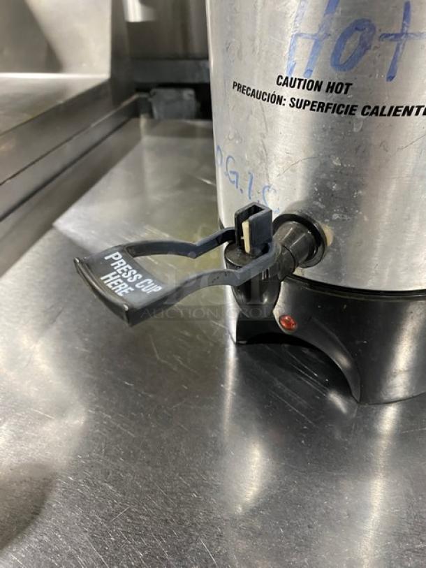 Stainless steel electric coffee urn with insulated design, spigot labeled "Press Cup Here," caution hot warning in English and Spanish.