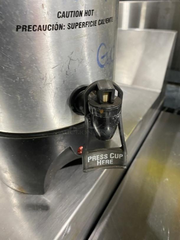 Commercial stainless steel coffee urn with "Caution Hot" label, press button dispenser, and insulated design. Used condition.
