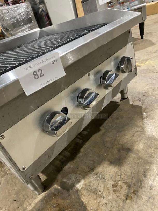 Commercial stainless steel gas countertop char broiler with wavy grates and legs, four control knobs, tag reading '82'.
