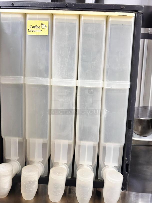 Five white plastic liquid dispensers with spouts labeled "Coffee Creamer" in a metal rack.