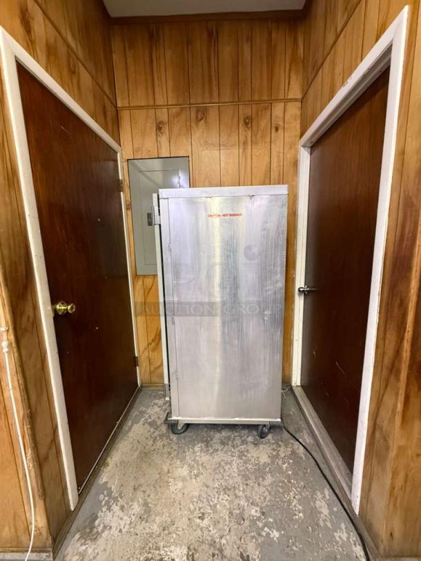 Full-size insulated proofer/warming cabinet, silver, on wheels, seen in a wood-paneled room. Markings indicate caution for hot surface.