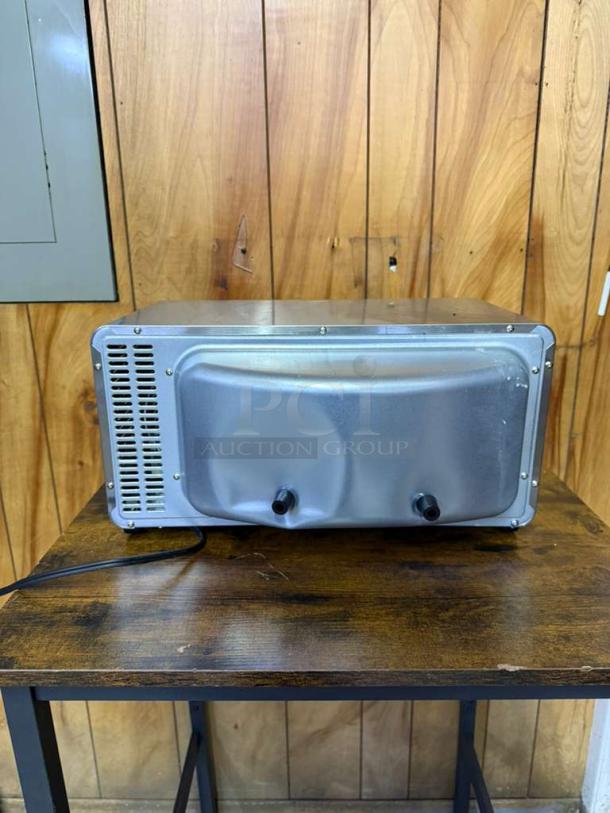 Back view of Hamilton Beach 4-Slice Toaster Oven, stainless steel, showing power cord and vent, on wooden surface.