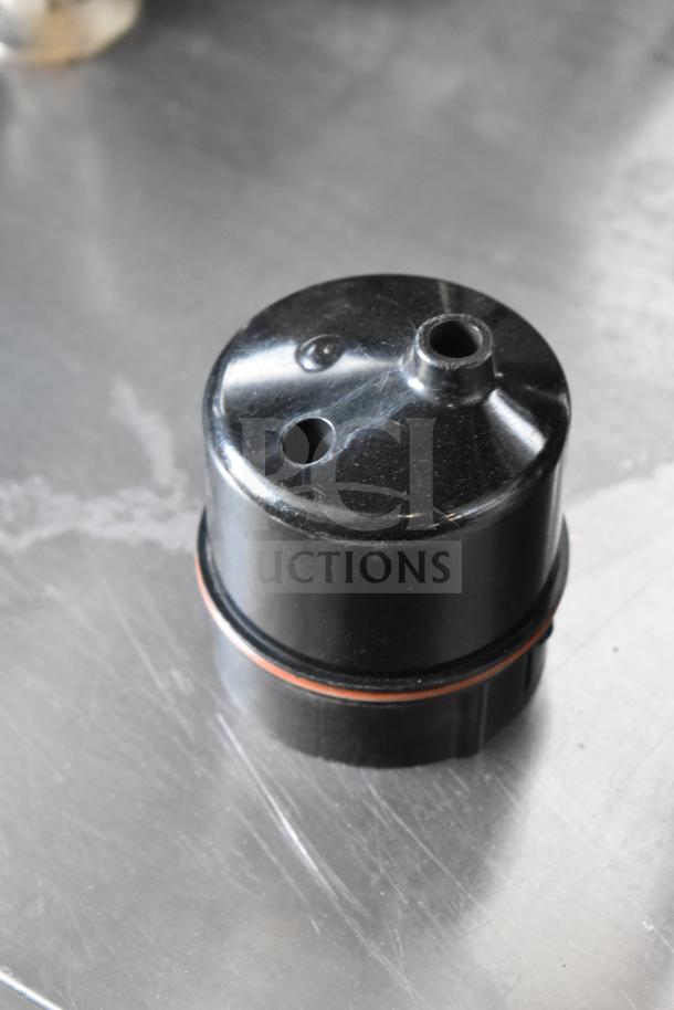 Black plastic component of Clover 1S coffee brewer, showing minor wear. Features two holes and an orange ring seal.
