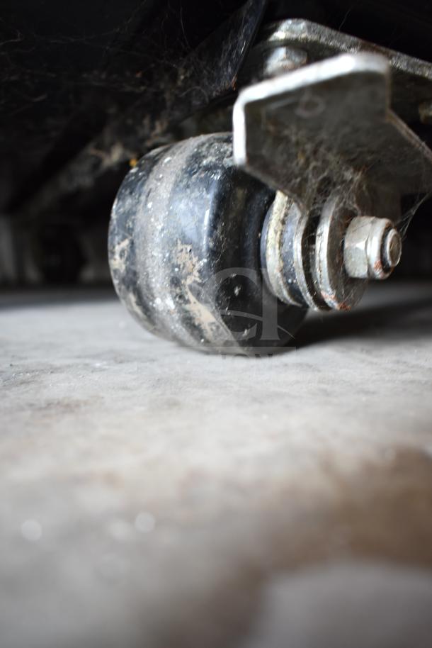 Close-up of a commercial caster wheel under the True TG1R-1S Reach In Cooler, showing wear and some dirt.