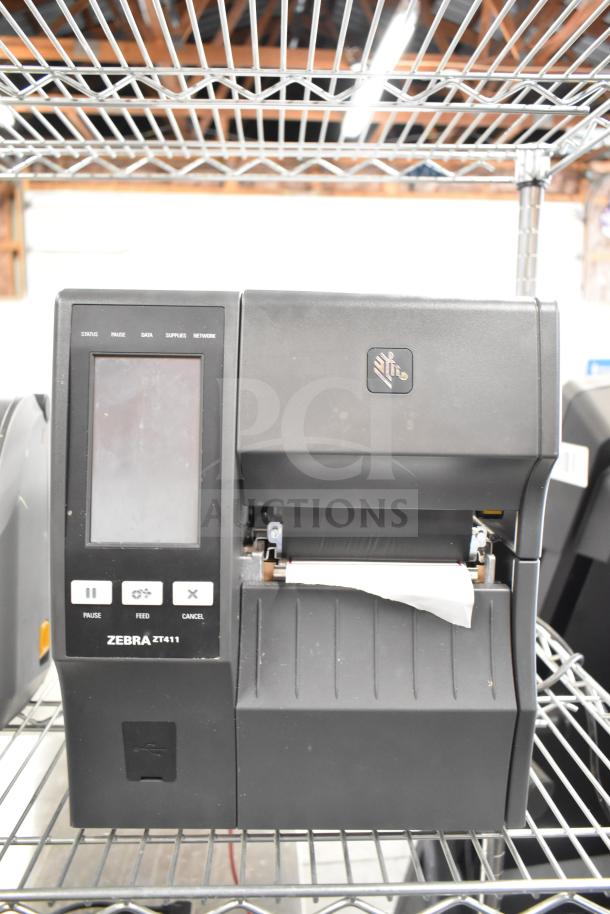 Zebra ZT411 Industrial Barcode Printer, black, with touchscreen, paper feed visible, used condition, on metal shelf.