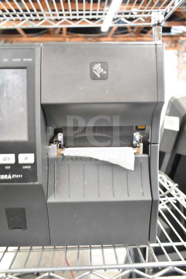 Zebra ZT411 Industrial Barcode Printer, black, used, visible paper output, frontal view on metal shelf.