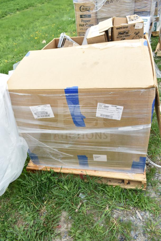 Large cardboard box wrapped in plastic on a wooden pallet, labeled with shipping barcodes and blue tape, outdoors.