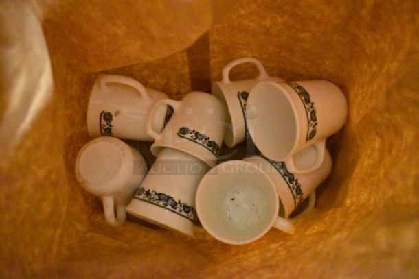 Eight white ceramic mugs with blue floral design, in used condition, stacked in a bag.