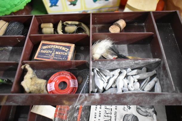 Vintage fishing tackle box with mixed contents including line, lures, sinkers, and an Independence safety matchbox.