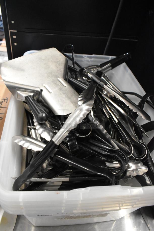 Lot of kitchen utensils in a clear poly bin, featuring metal tongs with black handles, in used condition.