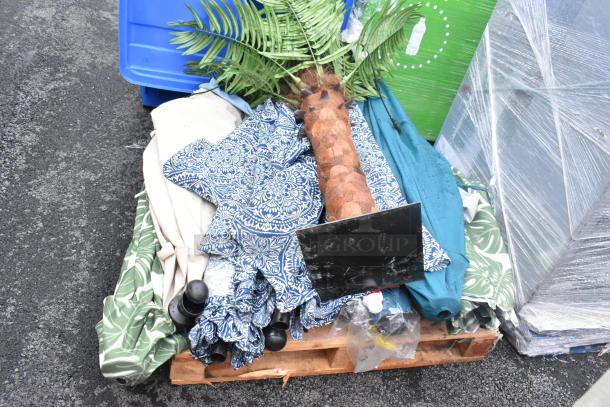 Assorted patio items including patterned umbrellas and artificial palm, bundled on a pallet. Various colors and patterns visible.