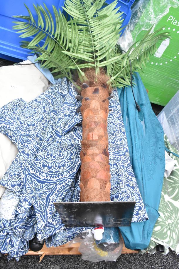 Assorted garden items with patio umbrellas, faux palm, and patterned blue fabric, laid on a pallet.