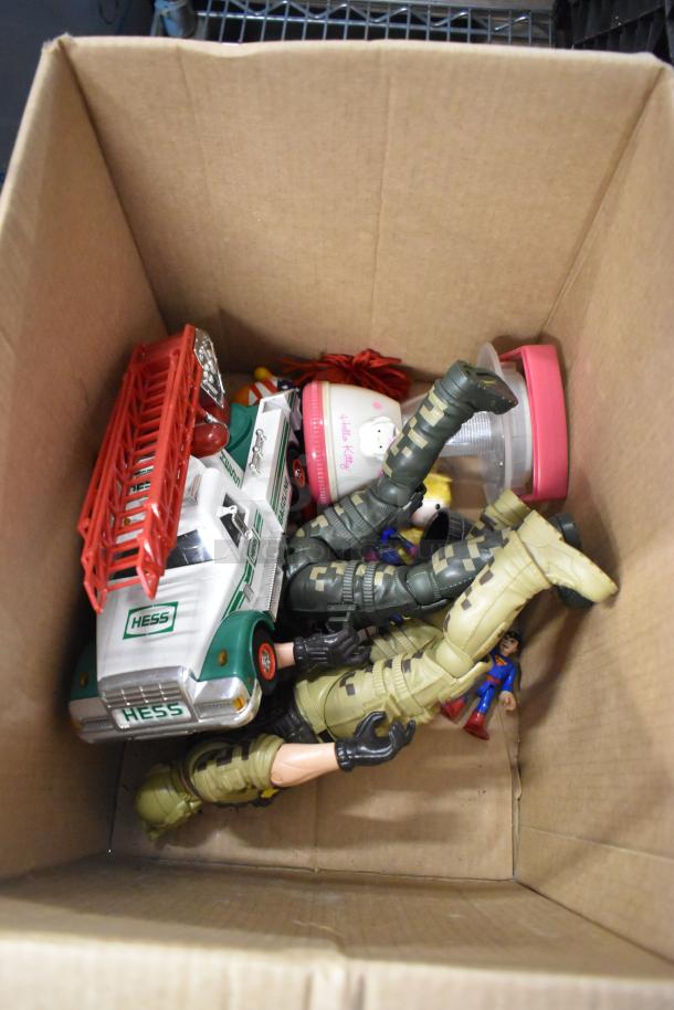 Cardboard box with Hess truck, various action figures, Hello Kitty cup, and toys. Mixed lot suitable for collectors.
