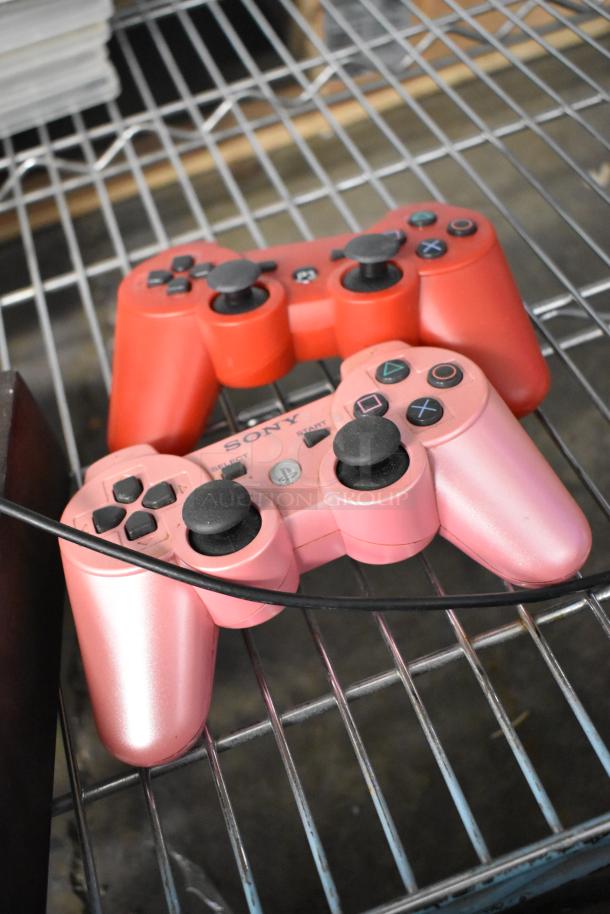 Two PS2 controllers, one pink and one red, on a metal shelf. Controllers show moderate wear.