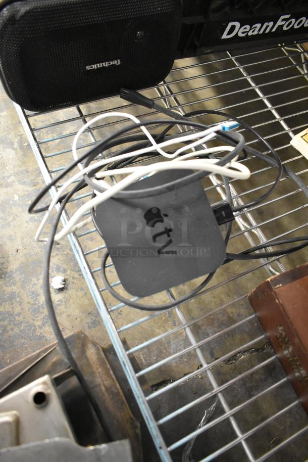 Mixed auction lot featuring a Technics speaker, Apple TV, and assorted cables on a metal shelf.
