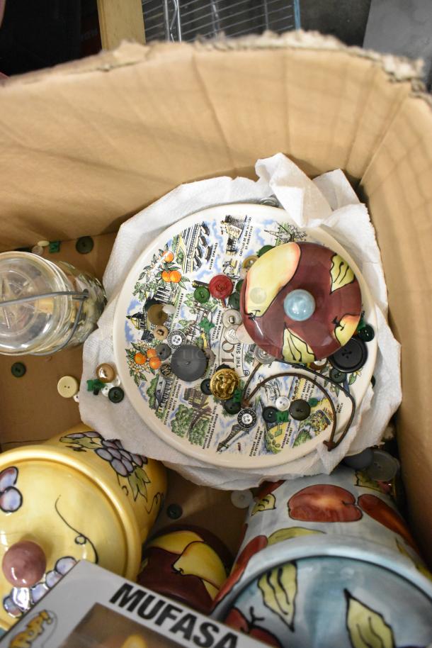 Assorted items in a box including a ceramic container with fruit design, state-themed plate, buttons, and Mufasa collectible.