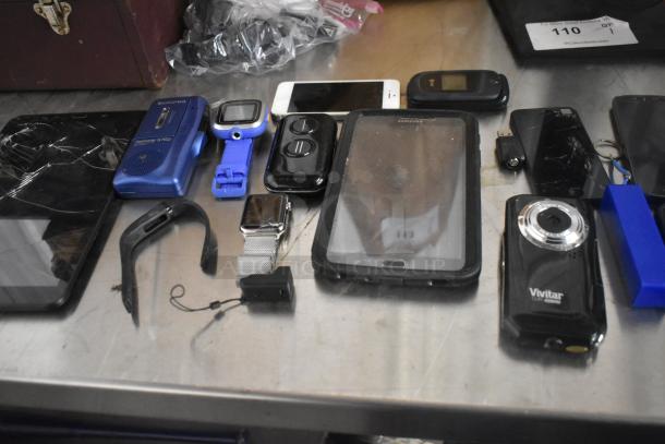 Assorted electronics including phones, tablets, smartwatches, and a Vivitar camera in various conditions on a table.