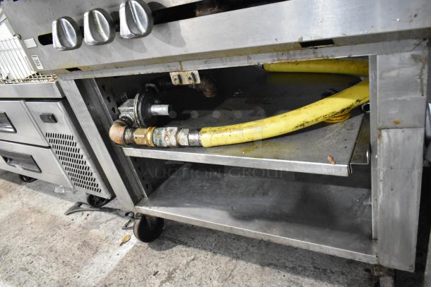 Stainless steel gas-powered French top range with knobs, two under shelves, and commercial casters. Visible gas line connections.