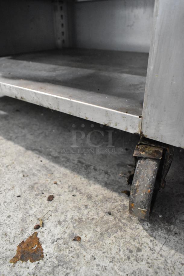 Stainless steel gas-powered French top range with single burner, two under shelves, on commercial casters, visibly worn.