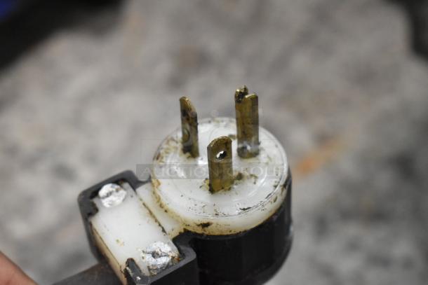 Close-up of a three-prong electrical plug, showing worn metal prongs and dusty surface, likely part of a 2021 Ampto SSGL Panini Press.