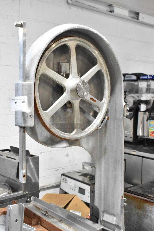 Metal commercial floor-style meat saw with large blade wheel, sturdy frame, and visible rust spots; auction tag present.