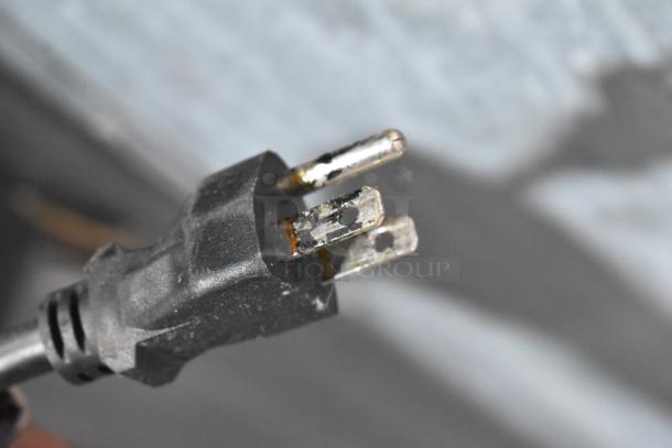 Close-up of a three-pronged plug with visible wear and rust on the metal prongs. Plug is attached to a black cord.