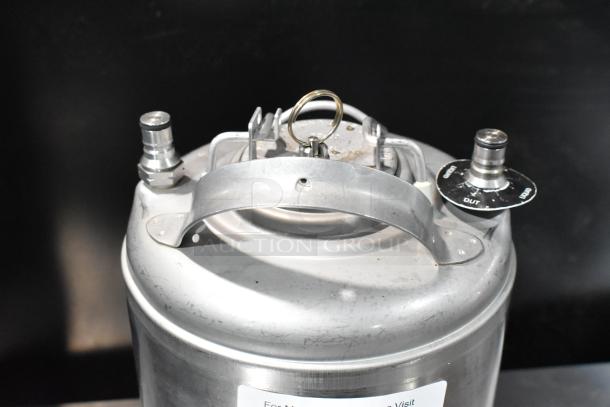 Stainless steel homebrew keg with dual valve connections, sturdy handle, and visible markings. Slightly scratched exterior.