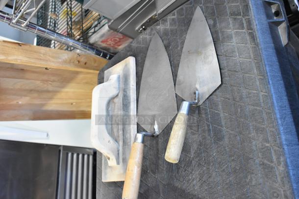 Two trowels and a hand float on a workbench, with two various bags containing contents, auctioned as 2 times your bid.