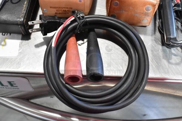 Coiled multimeter cables with red and black probes, lying on a metal table alongside leather and black cases.