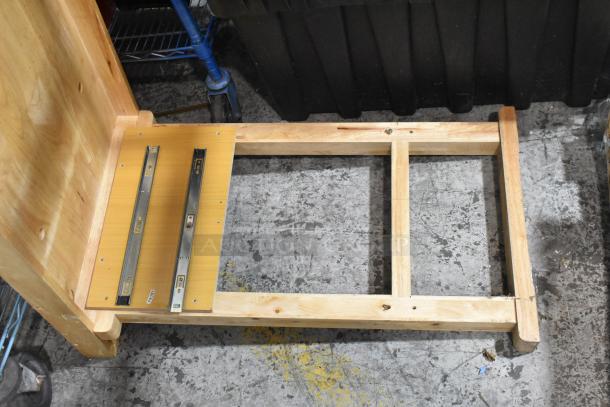 Wooden table base with unfinished light wood frame, metal brackets, and visible screws, on concrete floor. Balto label attached.
