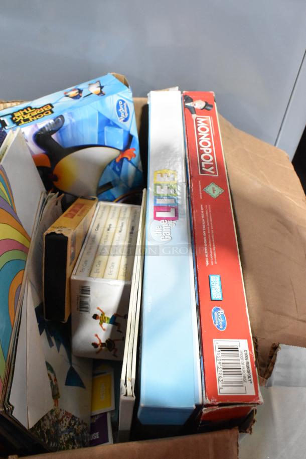 Cardboard box containing Monopoly and The Game of Life board games, mixed books, and toys. Items appear gently used.