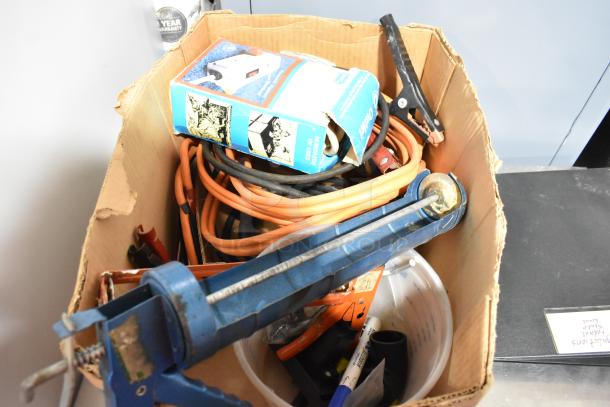 Box of assorted items including a blue caulk gun, orange jumper cables, and binders. Mixed condition, ideal for DIY projects.
