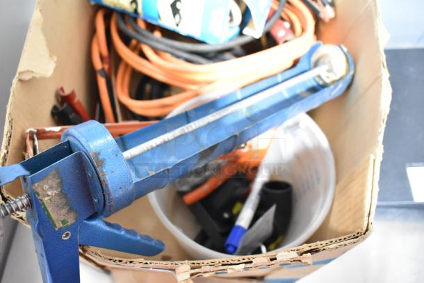 Cardboard box with blue caulk gun, orange jumper cables, binders, and various tools. Items show signs of wear.