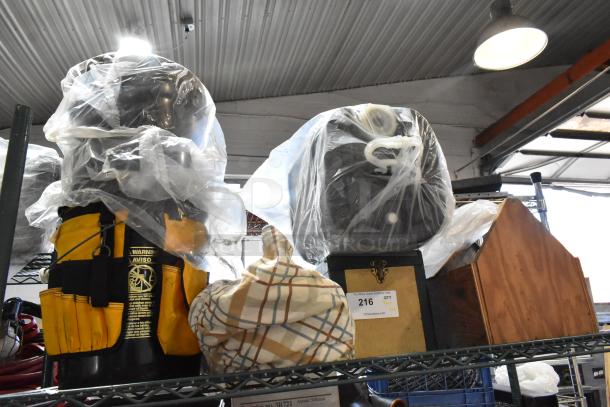 Alt text: Auction tier lot with a yellow tool bag, various plastic-wrapped items, plaid cloth, and a wooden box. Lot tag marked "216".