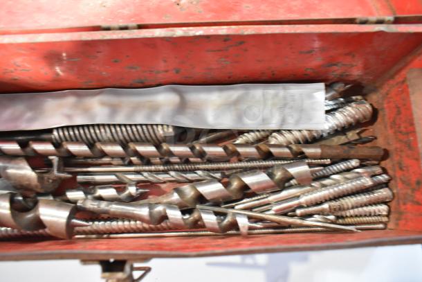 Red metal tool box containing various metal drill bits, showing signs of wear.
