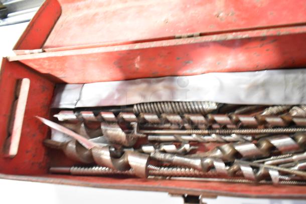 Red metal tool box containing assorted drill bits and tools, showing signs of wear. Ideal for collectors or DIY enthusiasts.