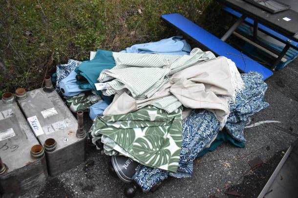 Pile of 18 various patio umbrellas with colorful patterns, including stripes and leaves. Some umbrellas have tags visible.