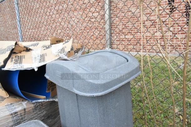 Four gray poly trash cans with lids, in good condition, nestled by a chain-link fence. Auction includes four units.