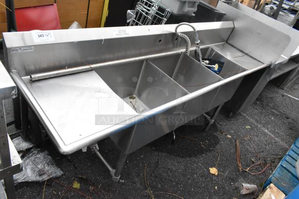 Stainless steel 3 bay sink with dual drain boards, featuring a gooseneck faucet. Condition appears used. Auction tags visible.