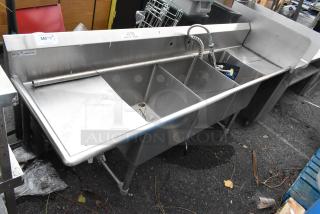 Stainless steel 3 bay sink with dual drain boards, featuring a gooseneck faucet. Condition appears used. Auction tags visible.