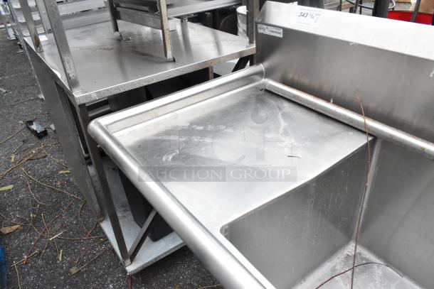 Stainless steel 3 bay sink with dual drain boards, showing slight wear. Features sturdy construction ideal for commercial use.