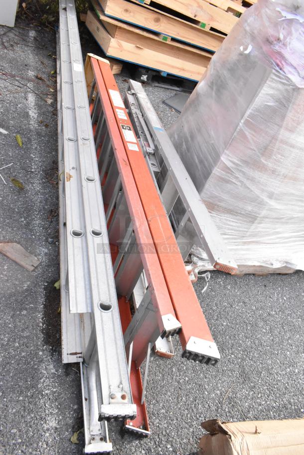 Three metal extension ladders, two silver and one orange, lying on the ground. Visible wear, labels intact.