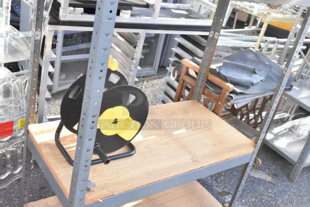 Gray metal shelving unit with wooden shelves, includes black and yellow hose reel on bottom shelf. Visible wear on surfaces.