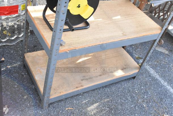 Gray metal shelving unit on asphalt surface, featuring two wooden tiers. Good condition, sturdy construction.
