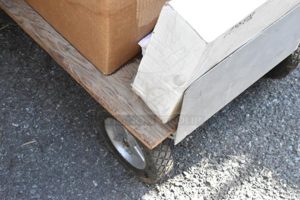 Flat cart carrying cardboard boxes, featuring sturdy wheels on pavement. Visible wood platform; functional condition.